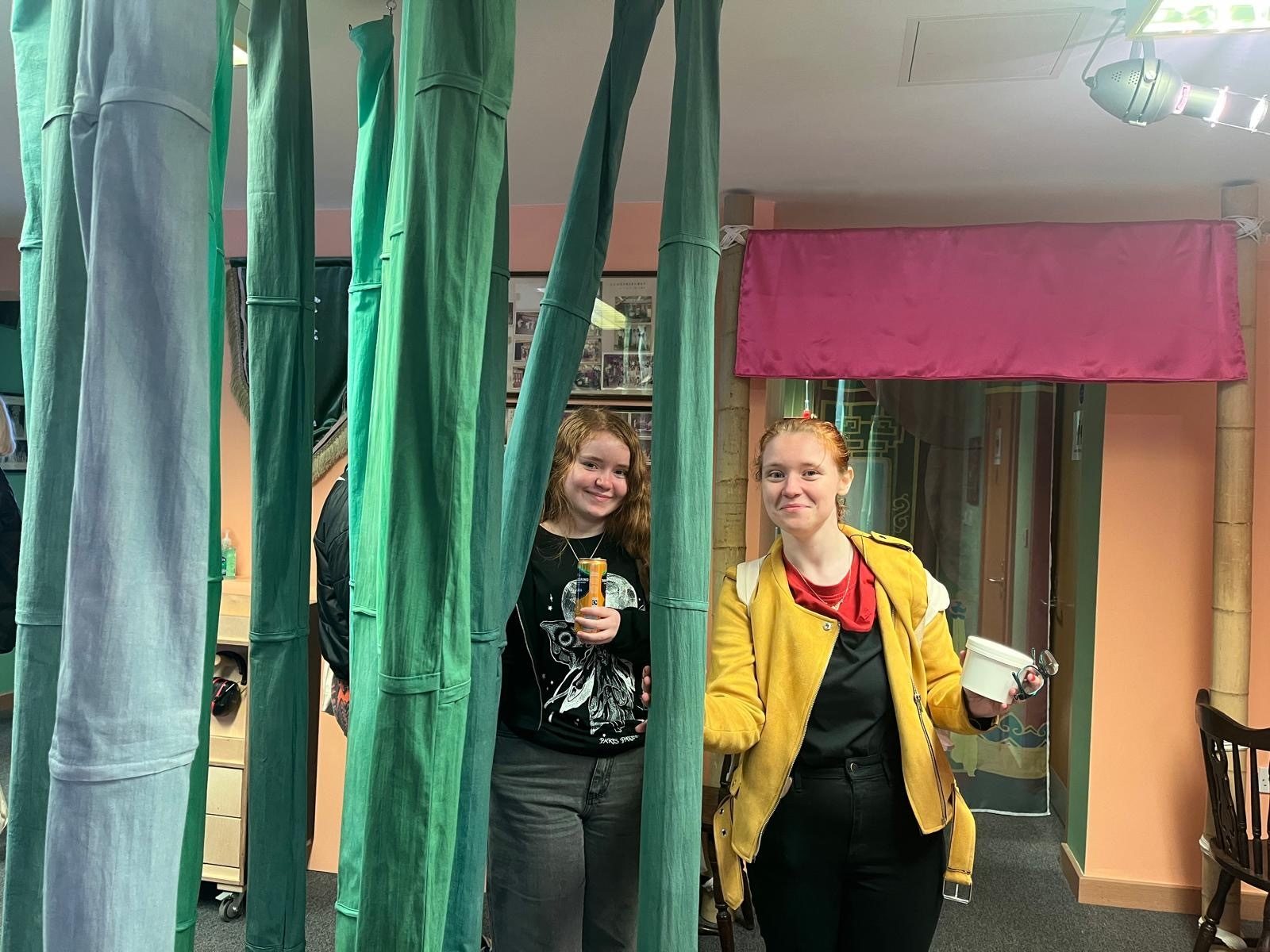 Two young women stand in a gallery setting, smiling at the camera through strands of hanging green fabric