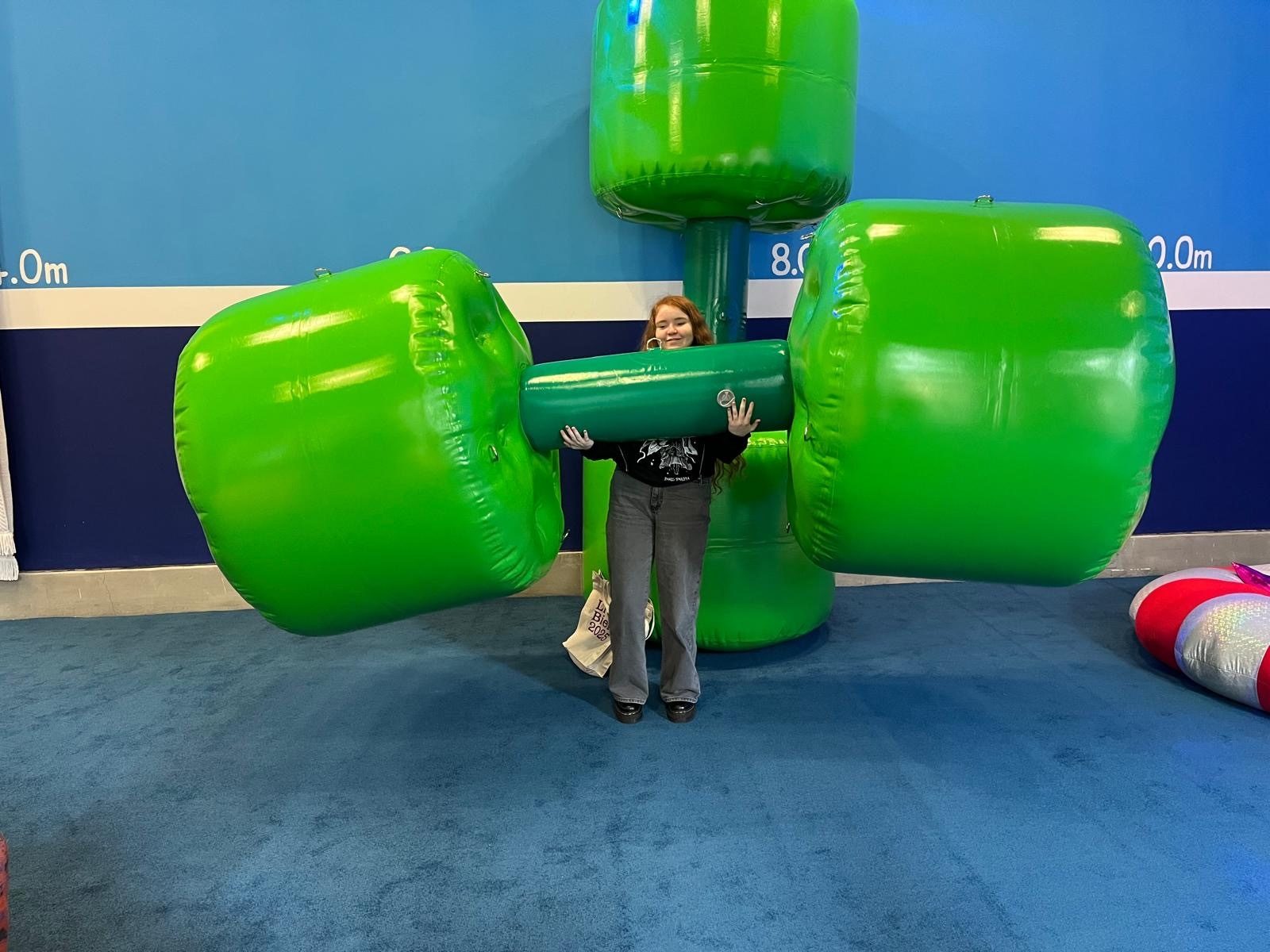 A young woman stands in a gallery setting holding up a giant inflatable dumbbell