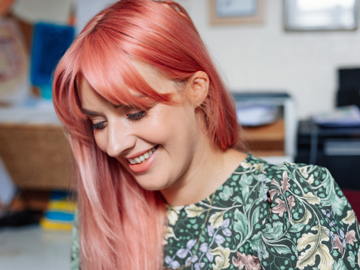 Photograph of a woman in her 20s/30s with long pink hair, wearing a green floral dress. She is smiling shyly