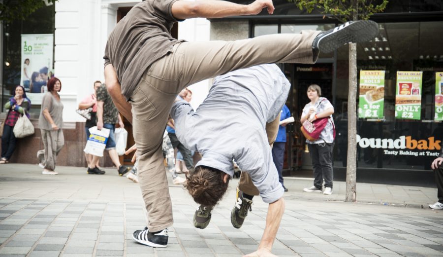 Two people dance on a high street. One is rolling towards the floor while the other swings their leg over the top of the other.