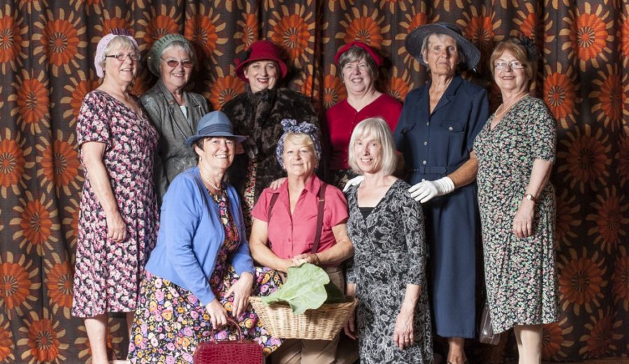 A group of people in vintage style floral dresses, and hats stand posed for a photograph in front of a colourful patterned curtain.