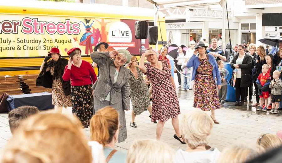 A group of people dressed in vintage style floral dresses, skirt suits and hats are dancing on a stage in front of an audience of people.