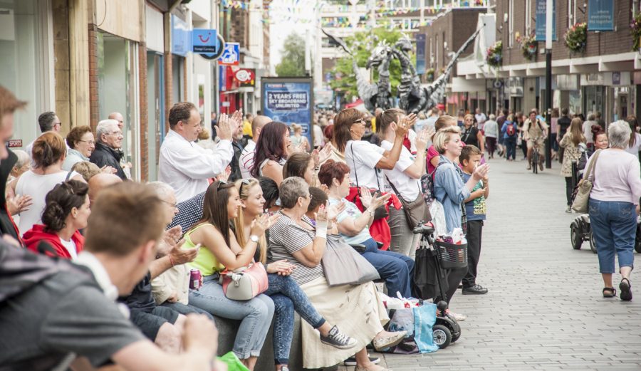 An audience of people sit and stand at the edge of a high street clapping.