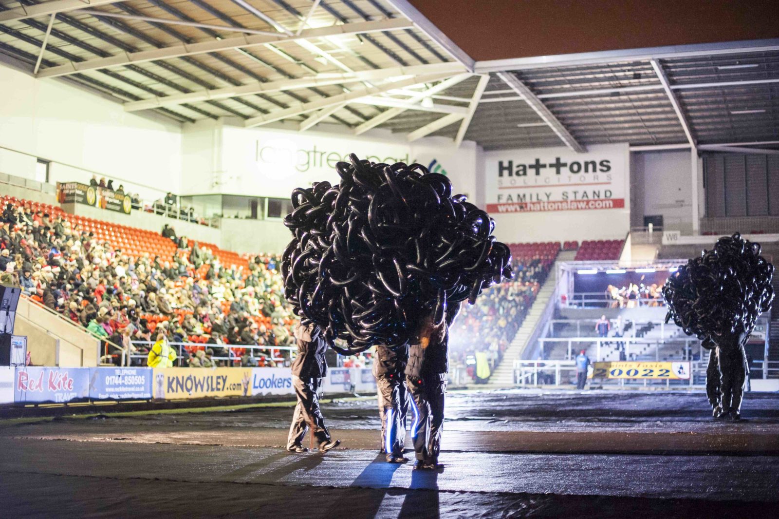 In a rugby stadium with an audience looking on two groups of people carry a large mass of black modelling balloons in a cloud shape around the pitch.