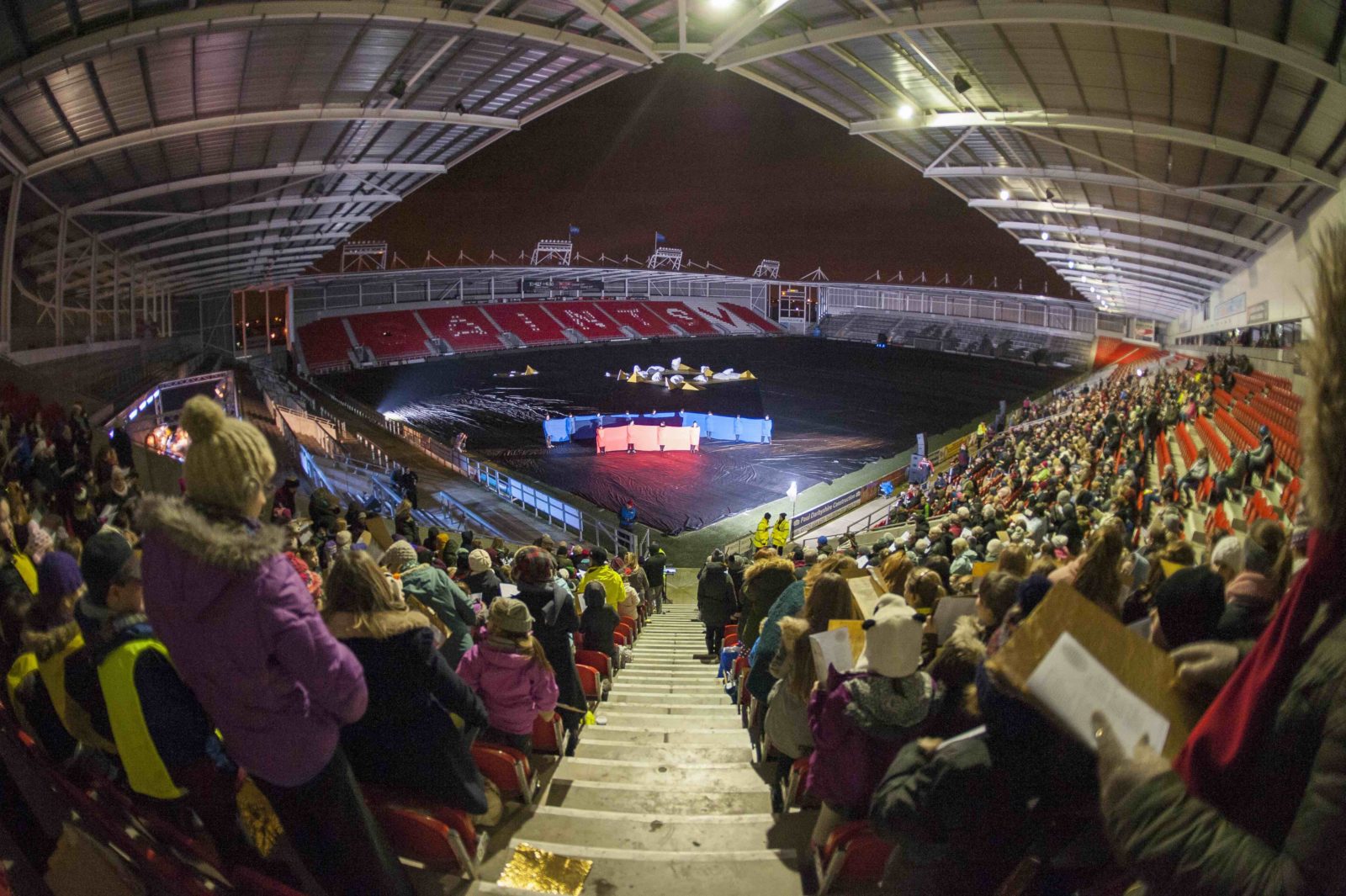 A rugby stadium with two of the seating areas full of people. We are looking down for the top of a corner set of stairs over the audience's shoulders towards the pitch, where a group of people stand wrapped in blue and red fabric.