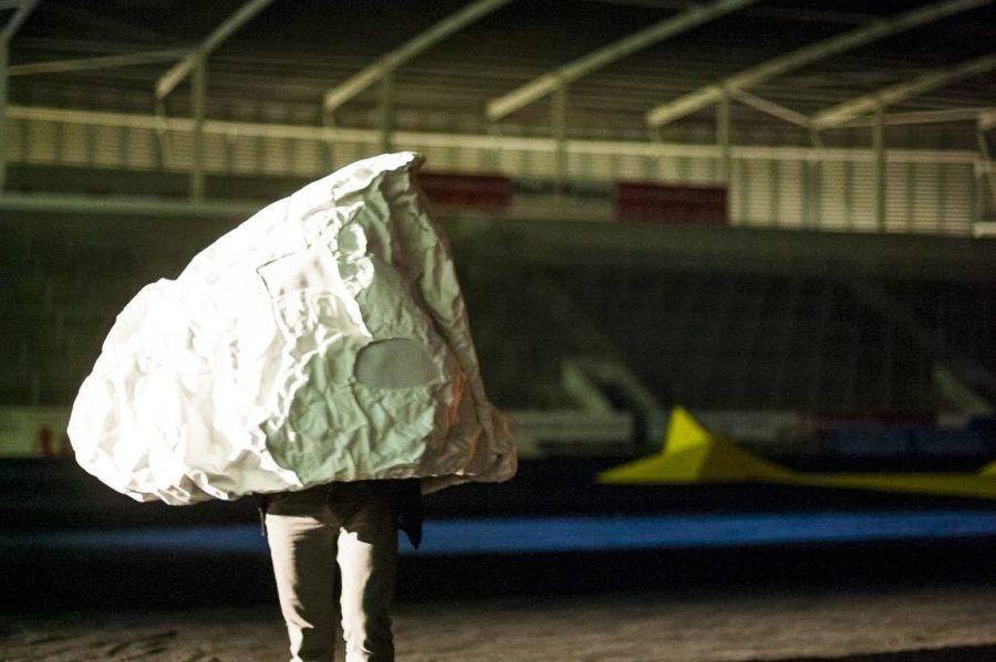 In a rugby stadium a large rock shaped sculpture has a person inside it with their legs visible. Under a night sky they stand on the pitch, which is covered by black material.