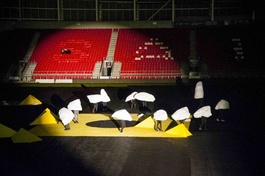In a rugby stadium, with empty red seats behind them, a collection of large rock shaped sculptures with human legs move around the pitch which is covered in black material and had yellow sculptures on the ground. They are lit by a spotlight.
