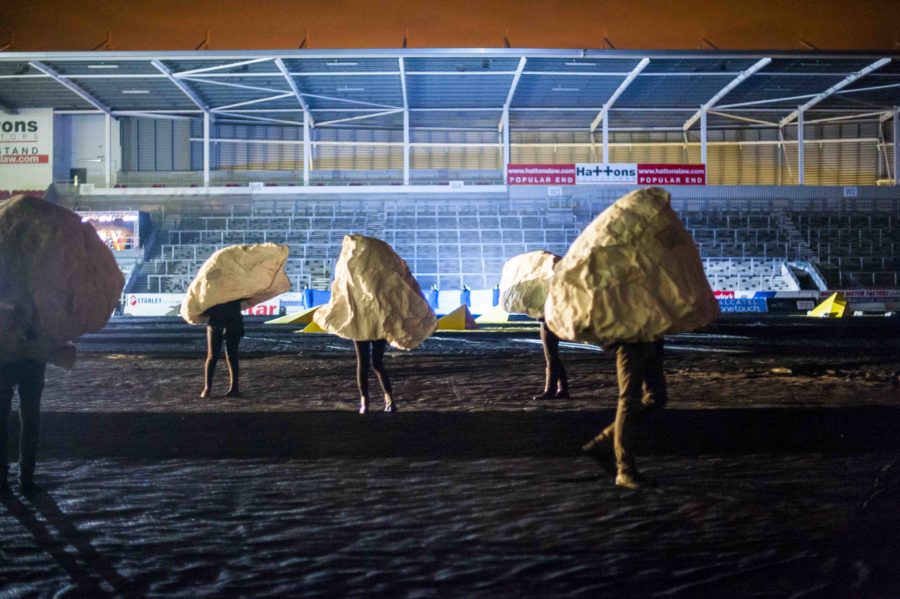 Five rock shaped sculptures with one person in each stand with their legs visible in an empty rugby stadium. It is night time and the pitch is lit by flood lights.