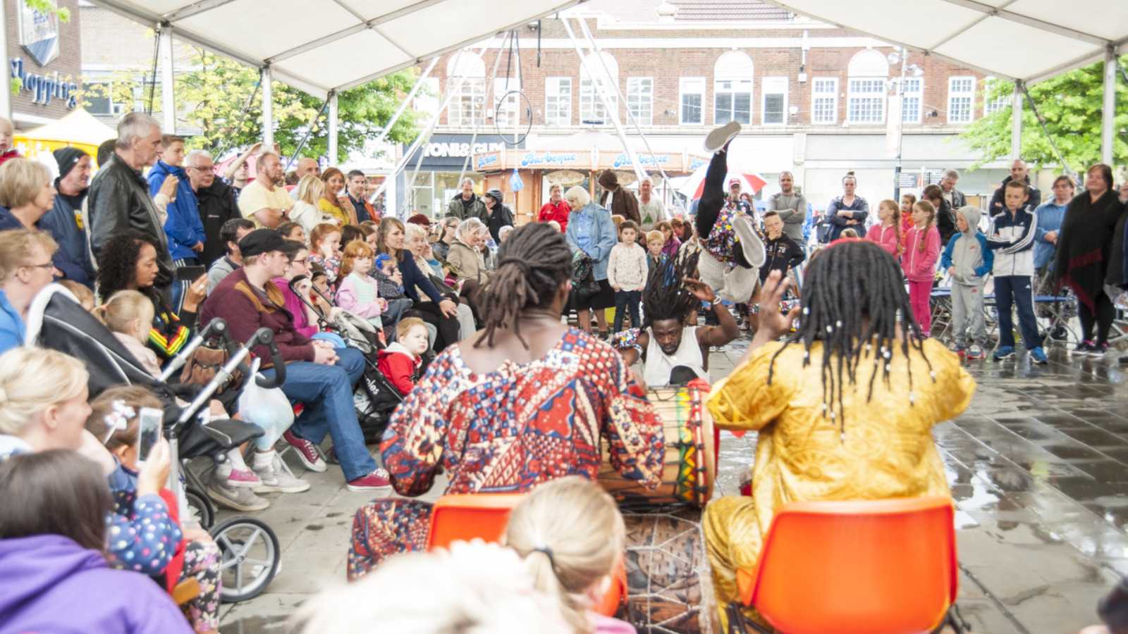 An audience of people sit and stand under a large gazebo in the middle of St Helens. Two people with dark, dreadlocked hair sit in orange seats ad play a pair of African drums while two people dance in front of them.