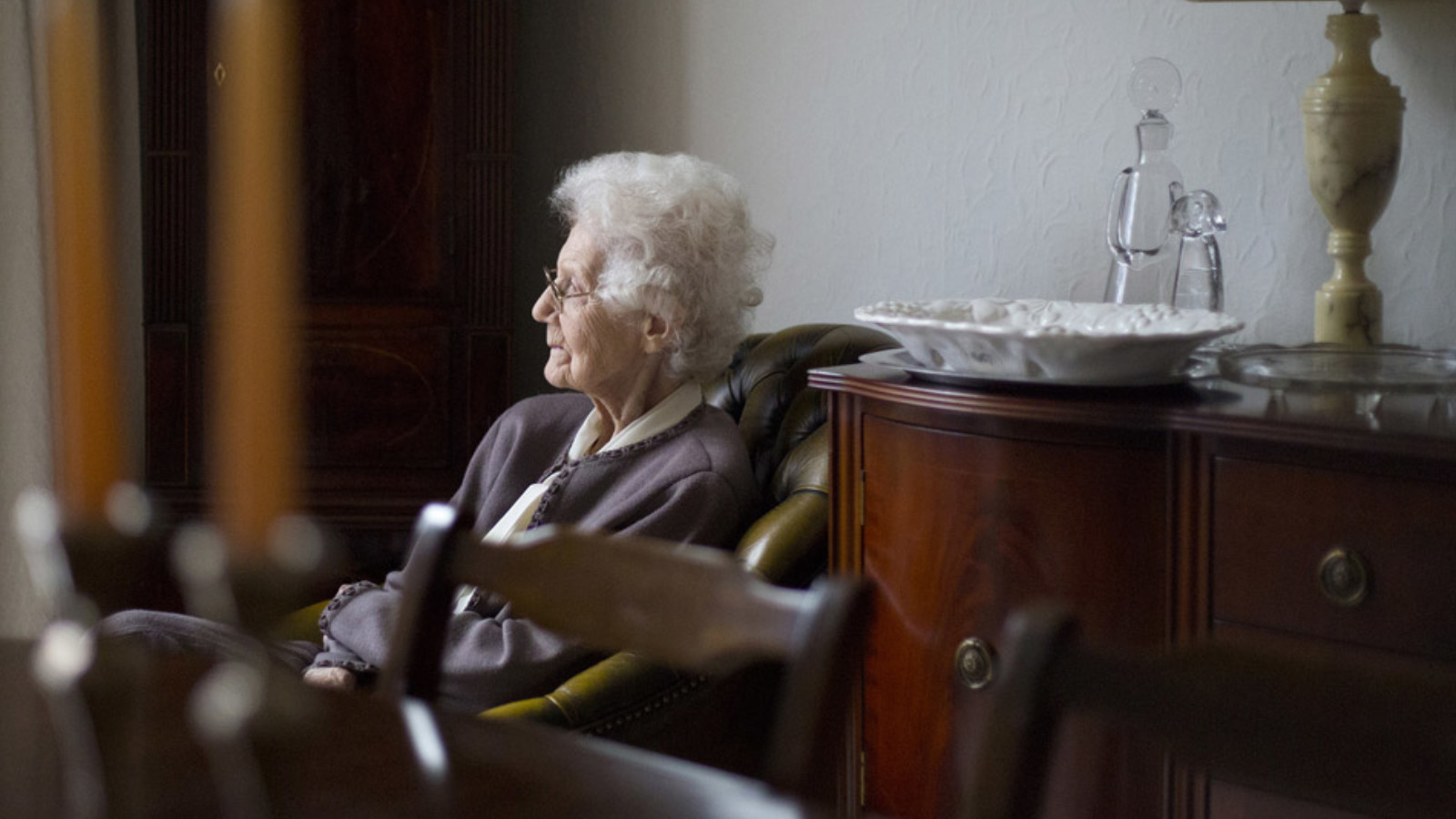 An older person with short, light hair sits in a room lit with daylight. They wear a plum jacket, white blouse and glasses. They sit on a leather chair next to a polished, dark wood sideboard with a lamp, decanter and plate on top. In the foreground of the image, slightly out of focus is a polished, dark wood dining table and chairs.