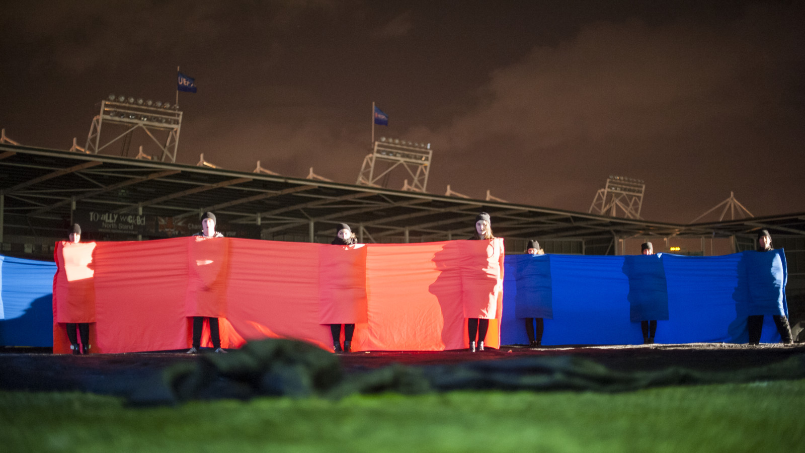 In a rugby stadium seven people stand on the grass under a night sky. They stand a meter away from each other and are connected by stretched out fabric, which also wraps around each of them. One group is wrapped in red fabric, another group in blue.