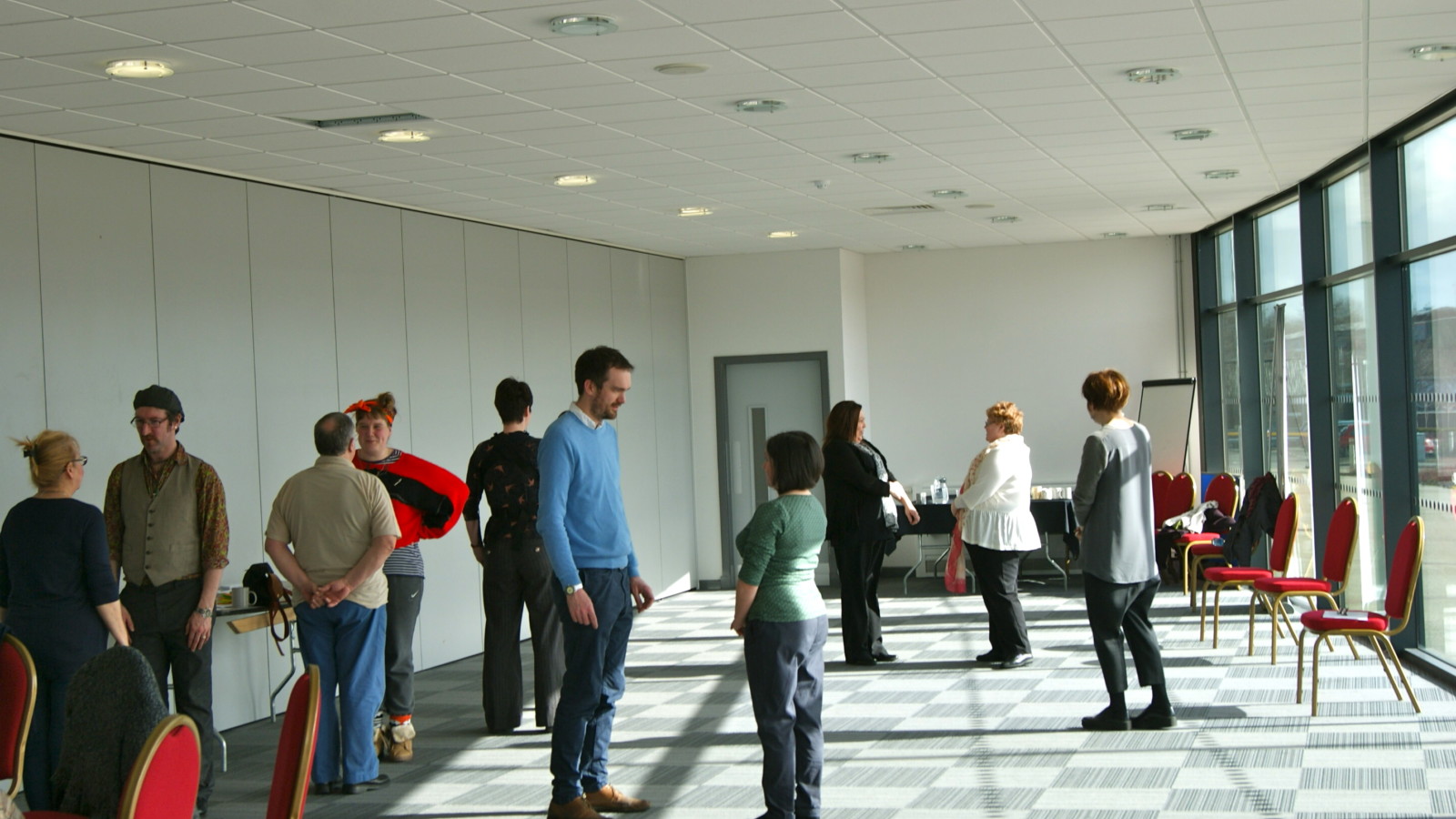 In a room lined all along one side with windows a number of people stand around the room in pairs. The pairs face each other standing around 1 meter apart.
