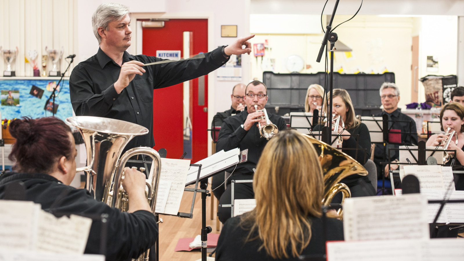 A person with short, light hair stands pointing with one hand and holding a conductor's baton in the other. They stand in a room in front of a number of people who are seated. The seated people are playing brass instruments. They have music stands with sheet music in front of them.