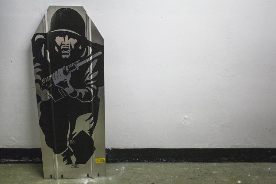 A piece of metal leans against a white wall. The metal is painted with a stencilled painting of a person in uniform charging with a gun.