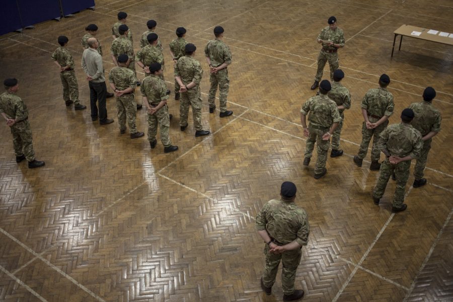 A group of people stand in army uniform in a large hall. They are being addressed by one person in uniform at the front.