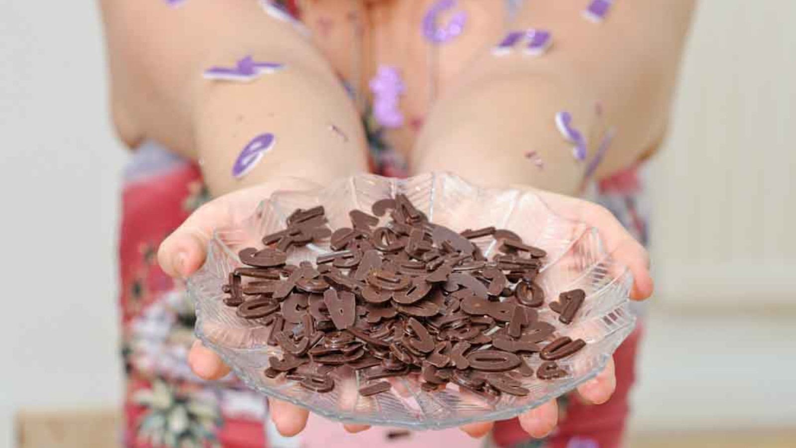 A pair of hands hold a glass dish holding chocolate letters. The persons arms have purple letters stuck to them.