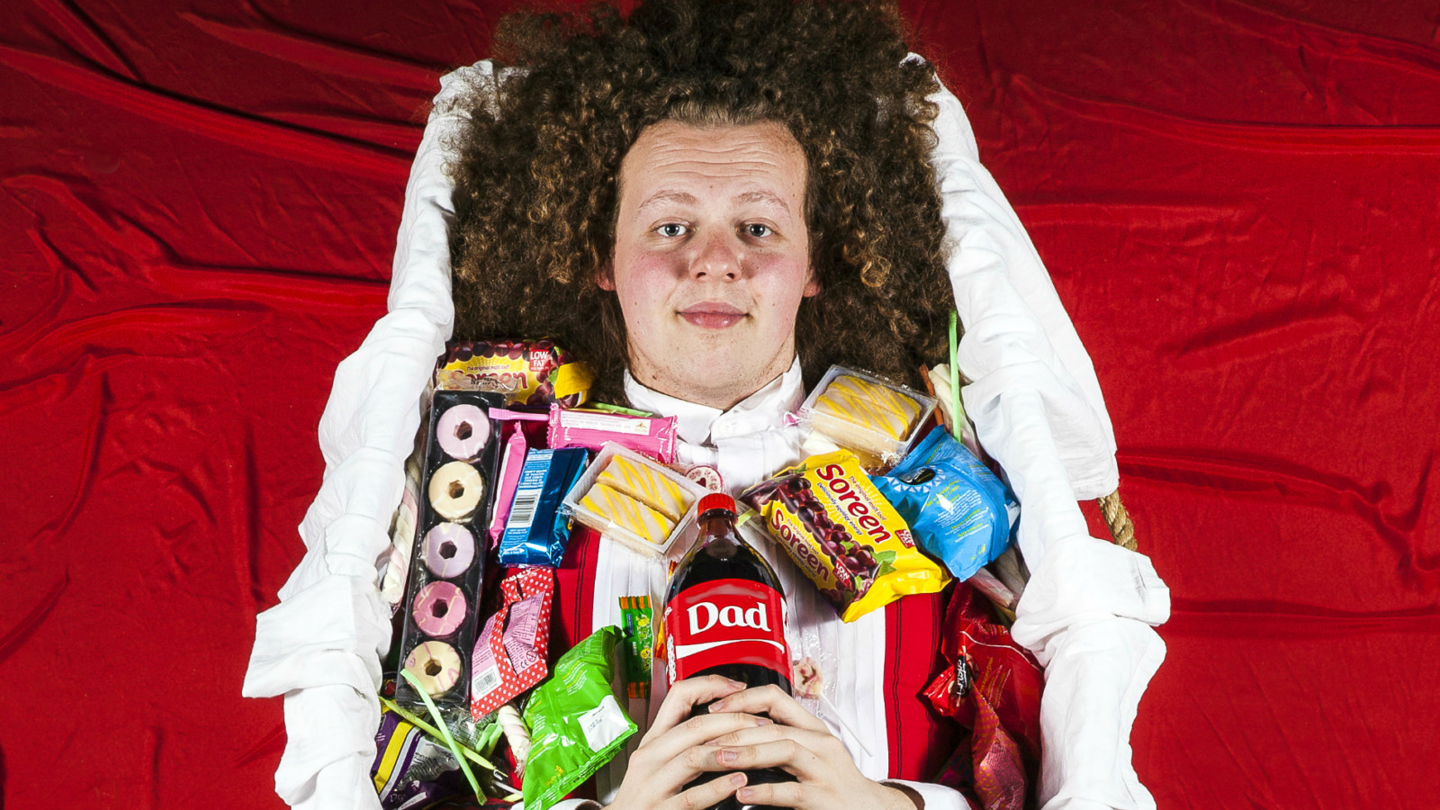 The artist Jack Rooke lies in a white lined coffin with various confectionary and holding a Coca Cola bottle with 'Dad' on the label.