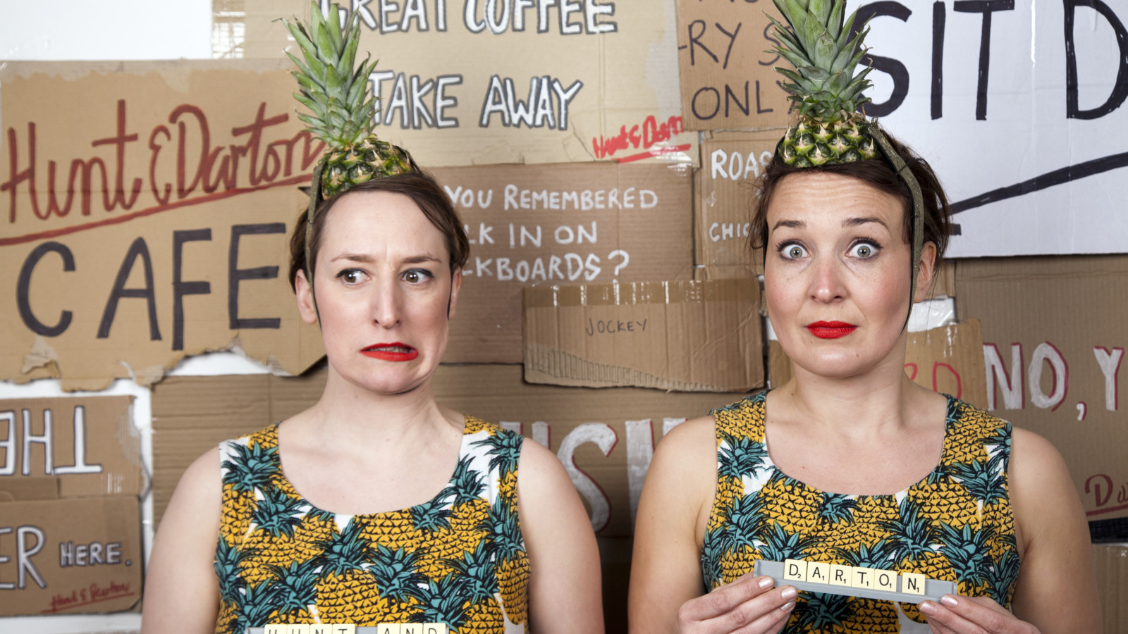 Artists Jenny Hunt and Holly Darton wear pineapple patterned clothes and pineapple tops on their heads. Their faces are surprised. They hold scrabble pieces which say 'Hunt and Darton' and are standing against a backdrop of many cardboard signs.
