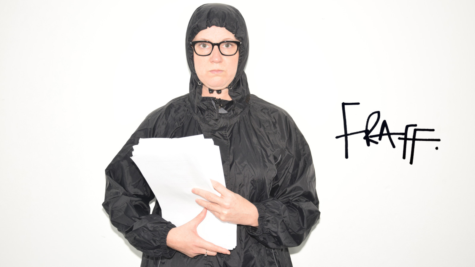 The artist Barbara Brownskirt stands in a black anorak with the hood up and holding a stack of white papers against a white background. The word 'FRAFF' is written on the background.