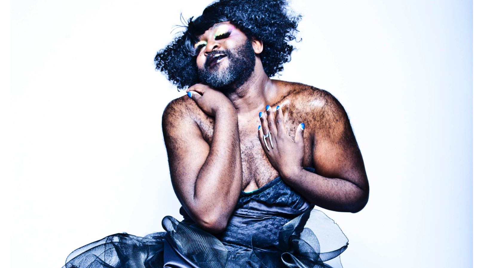 The artist Le Gateau Chocolat stands against a white background. They wear a black cocktail dress, blue nail varnish and curly hair. They have their eyes closed, smiling and are touching their chest.