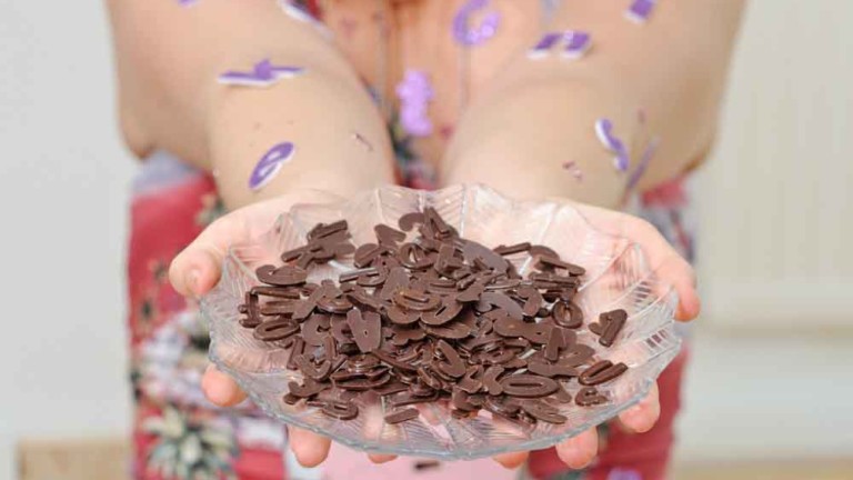 A pair of hands hold a glass dish holding chocolate letters. The persons arms have purple letters stuck to them.