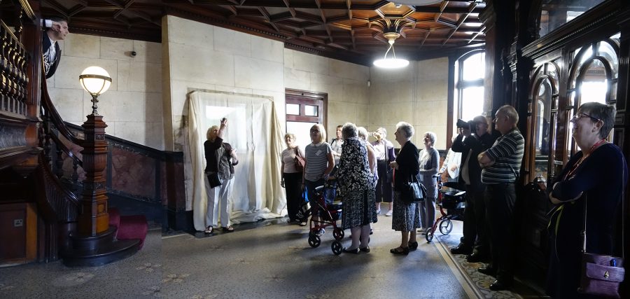 A group of people stand in a room with ornate tiled floors, dark wood panelled ceilings and large ornate doors. Many of the people in the group are smiling and looking towards a staircase. Two people are taking photographs. On the left the large, dark wood staircase.