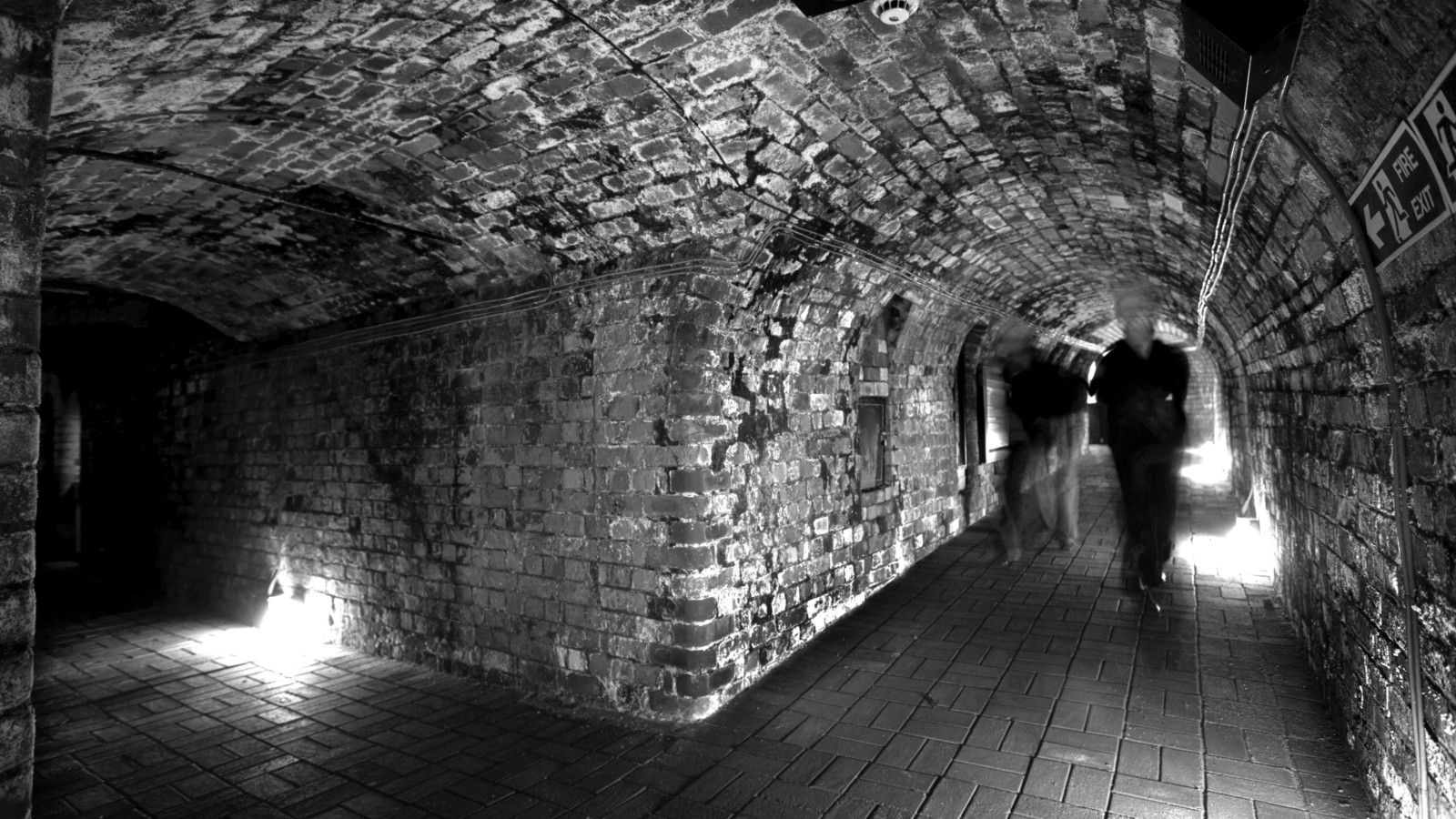 A black and white photograph of dark, brick tunnels lit by a small amount of lights at ground level. Two blurred figures are walking through the tunnels.