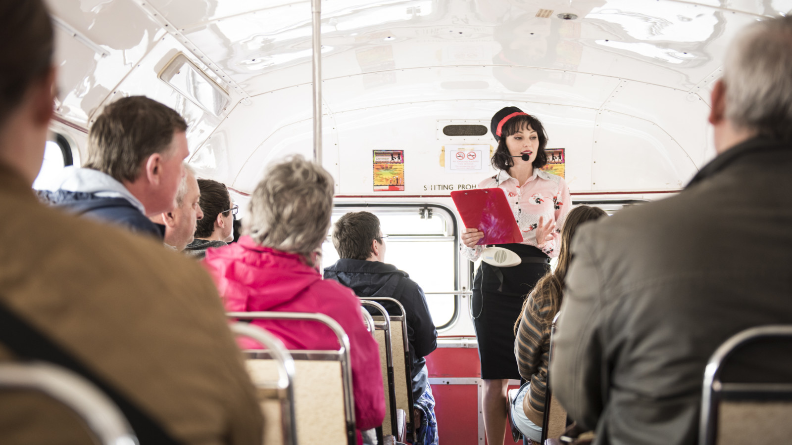 The artists Lowri Evan stands in a vintage style outfit wearing a headset and holding a clipboard on the top deck of a bus. talking to an audience of people sitting in the bus seats
