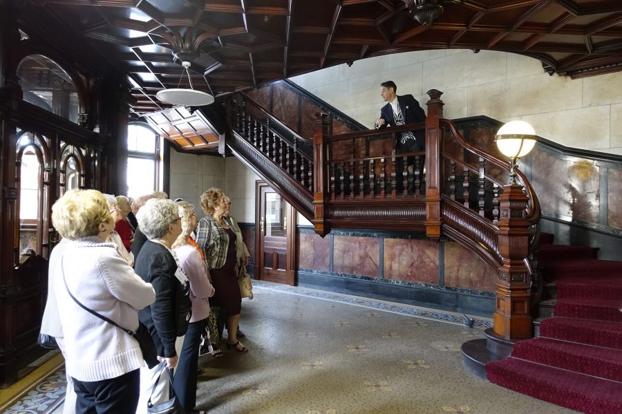 A group of people stand in a room ornate tiles floors and walls and a dark wood panelled ceiling. In the room is also a large, dark wood staircase with dark red carpet. On the staircase a man in a dark suit leans over the rail talking to the group of people.