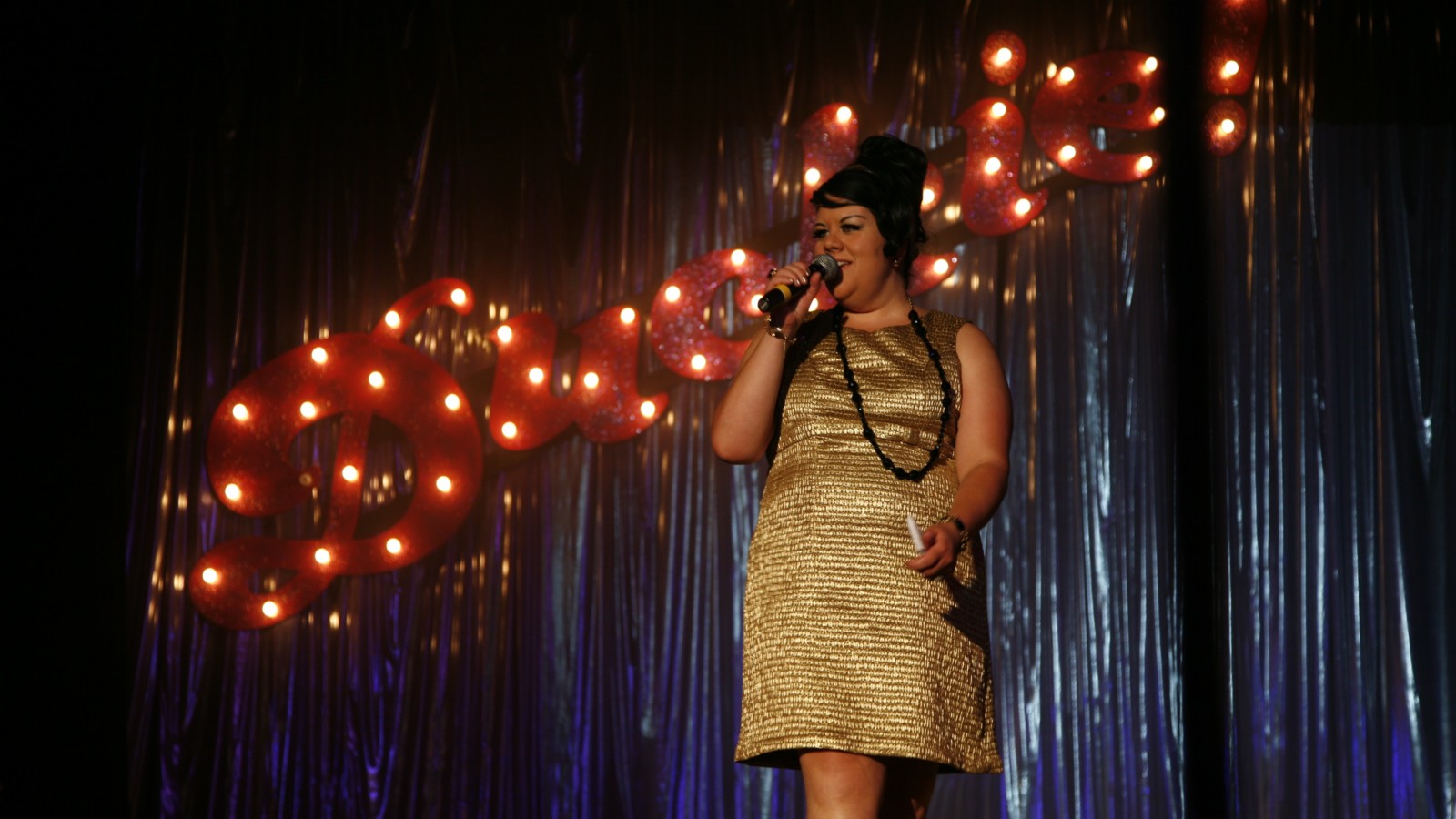 Artist Amy Lame stands in a gold dress holding a microphone in front of a metallic blue curtain and the word 'Duckie' in large red, letters with lights.