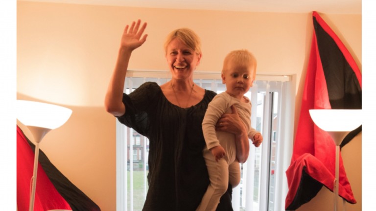 A person with blonde hairs stands in a room lit with lamps and some natural light form a window behind them. They hold a small child a baby grow and are waving.