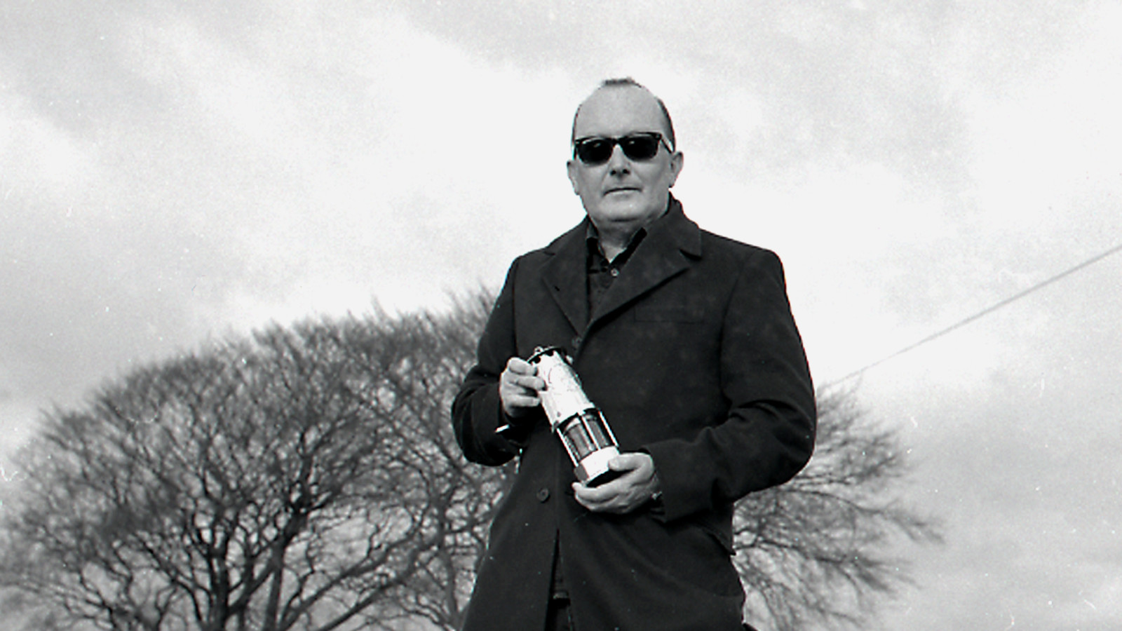 A black and white image of a man in a dark coat and sunglasses holding a miners lamp.