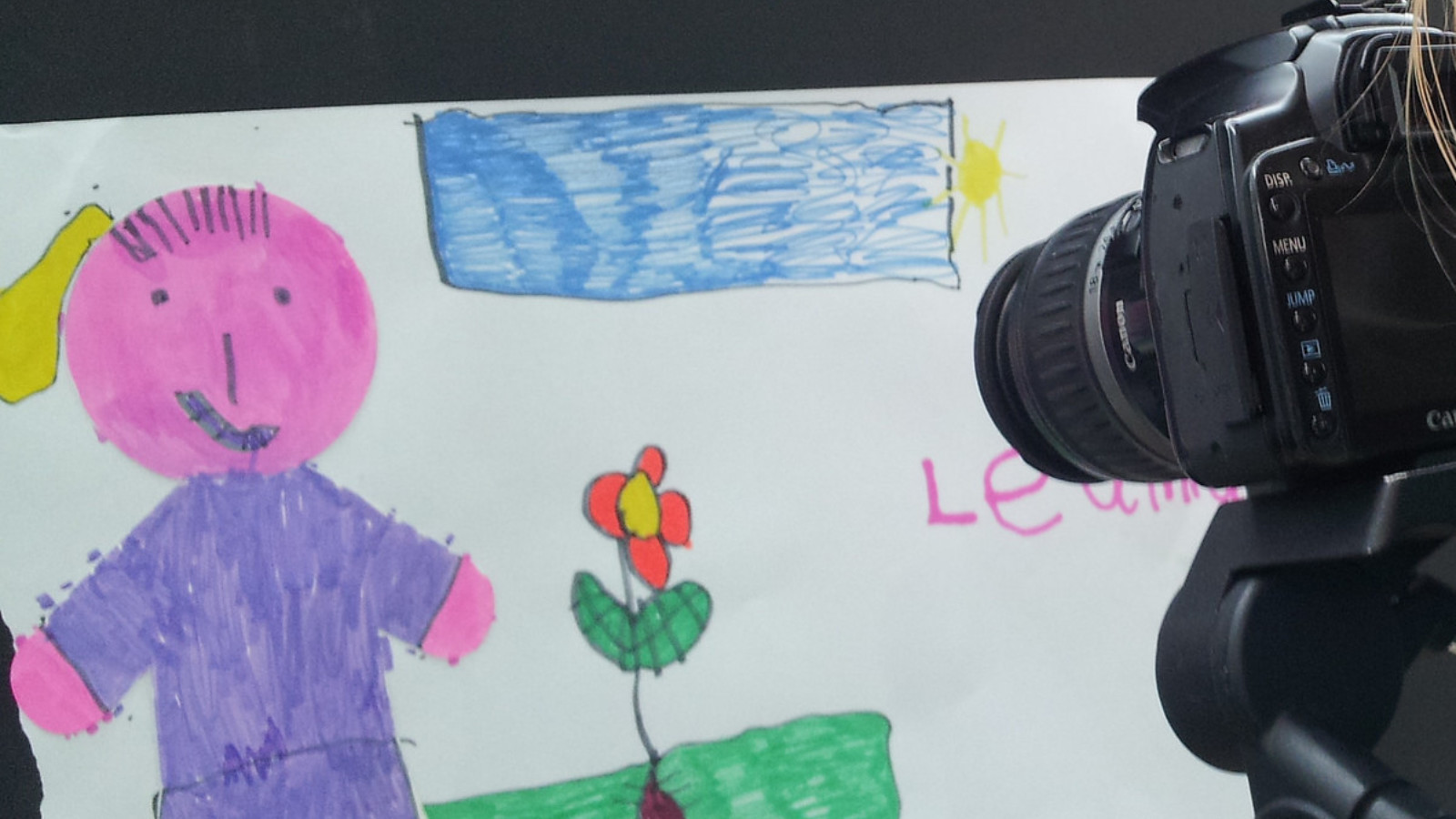 A digital camera is pointed towards a felt tip drawing of a person standing on a patch of grass next to a flower.