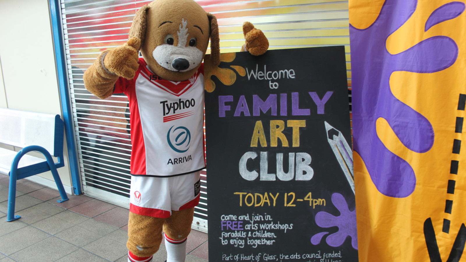 A mascot which is a dog dressed in a white and red rugby kit stands with two thumbs up in front of a window and next to a blackboard sign. The sign reads Welcome to Family Art Club