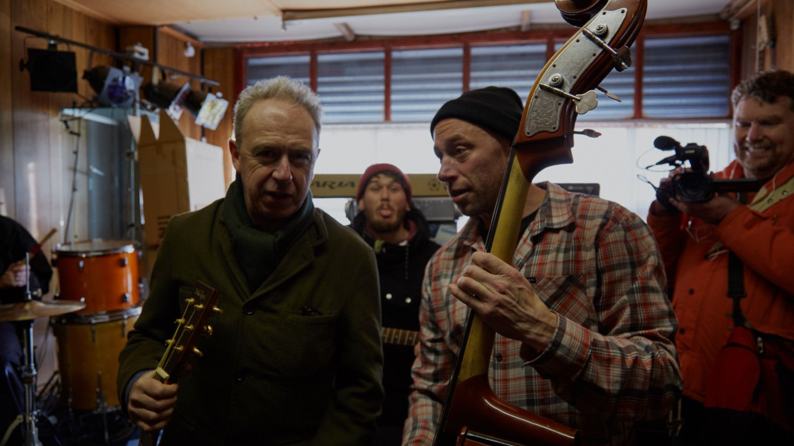 Four people stand in a wooden panelled room with a large window behind them. There is a drumkit and a set of disco lights to the left. Two of the people stand in the forefront. One, with short light hair and wearing a dark coat holds a an acoustic guitar by the neck. The other, wearing a black woollen hat and a checked shirt holds a double bass. In the background a person in a dark red hat pulls and tongue and another wearing a red coat points a video camera towards the musicians and smiles.