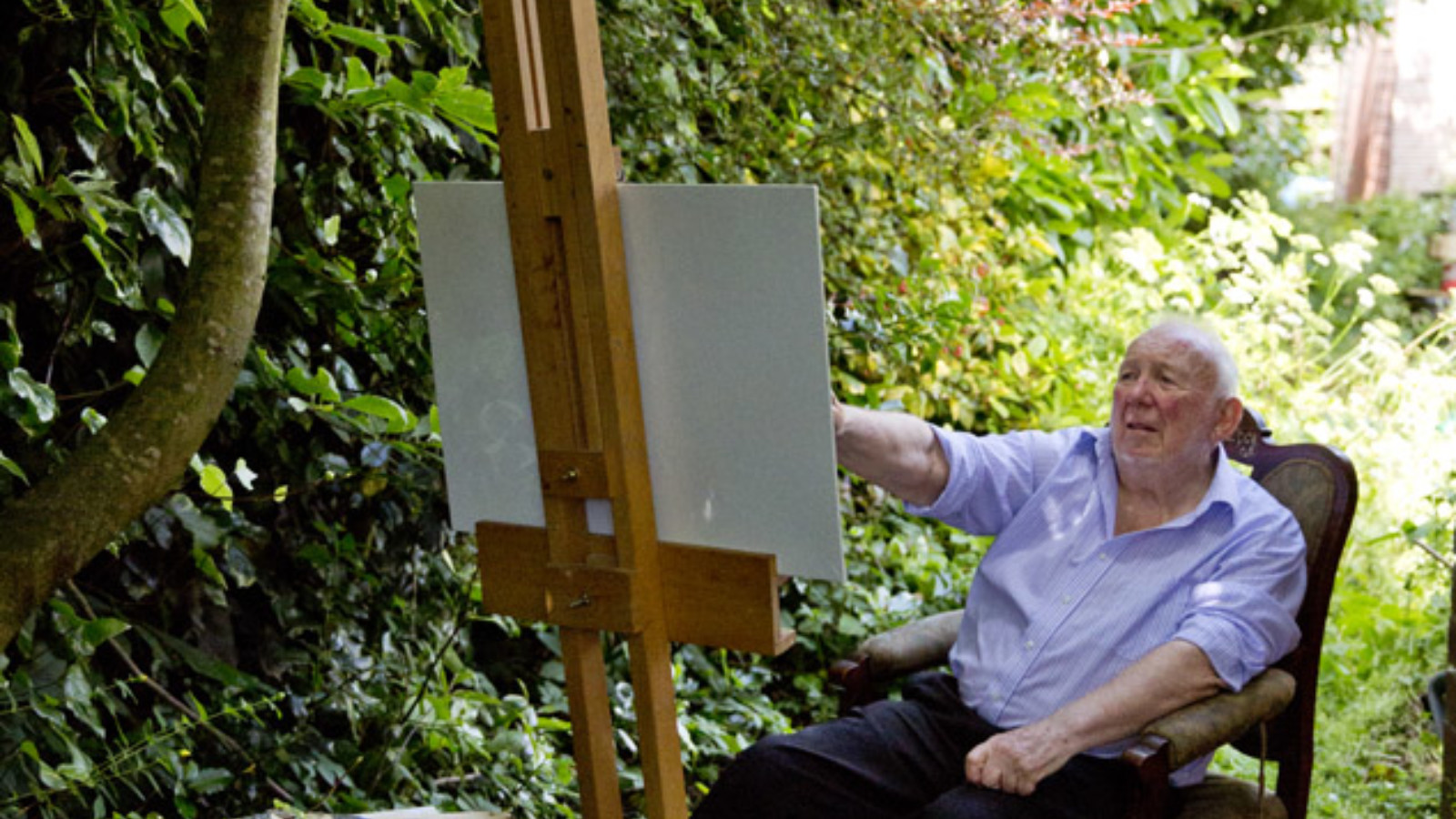 An artist sits in a chair in front of an easel and surrounded by arts materials. They are sitting outside, in daylight in a tunnel of trees.