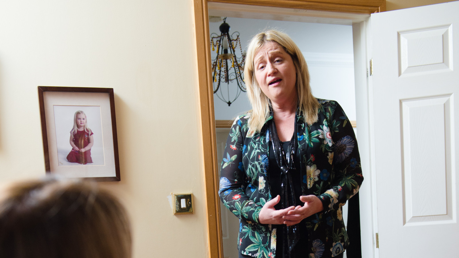 A woman with light hair and a patterned blouse stands in a doorway inside a house living room. She is singing. We can see in the corner the back of a persons head,  they are watching the woman sing.