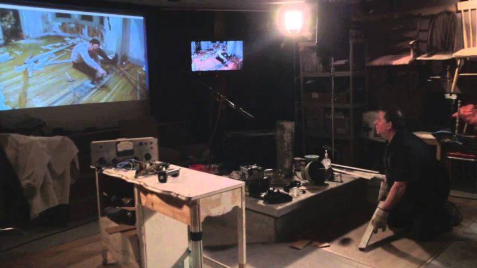 An image of a person kneeling in a dark room, lit by one lamp. They are surrounded by camera and other visual imagery kit and are looking at a projector screen and TV with an image of a person sitting on a wooden floor.