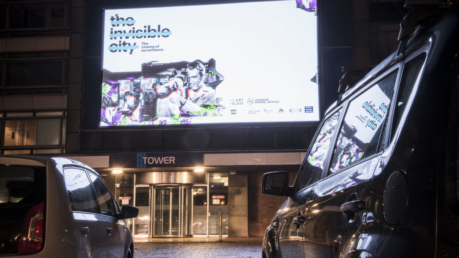 An LED billboard on a tower block displaying a graphic reading 'The Invisible City: The City of Surveillance'.