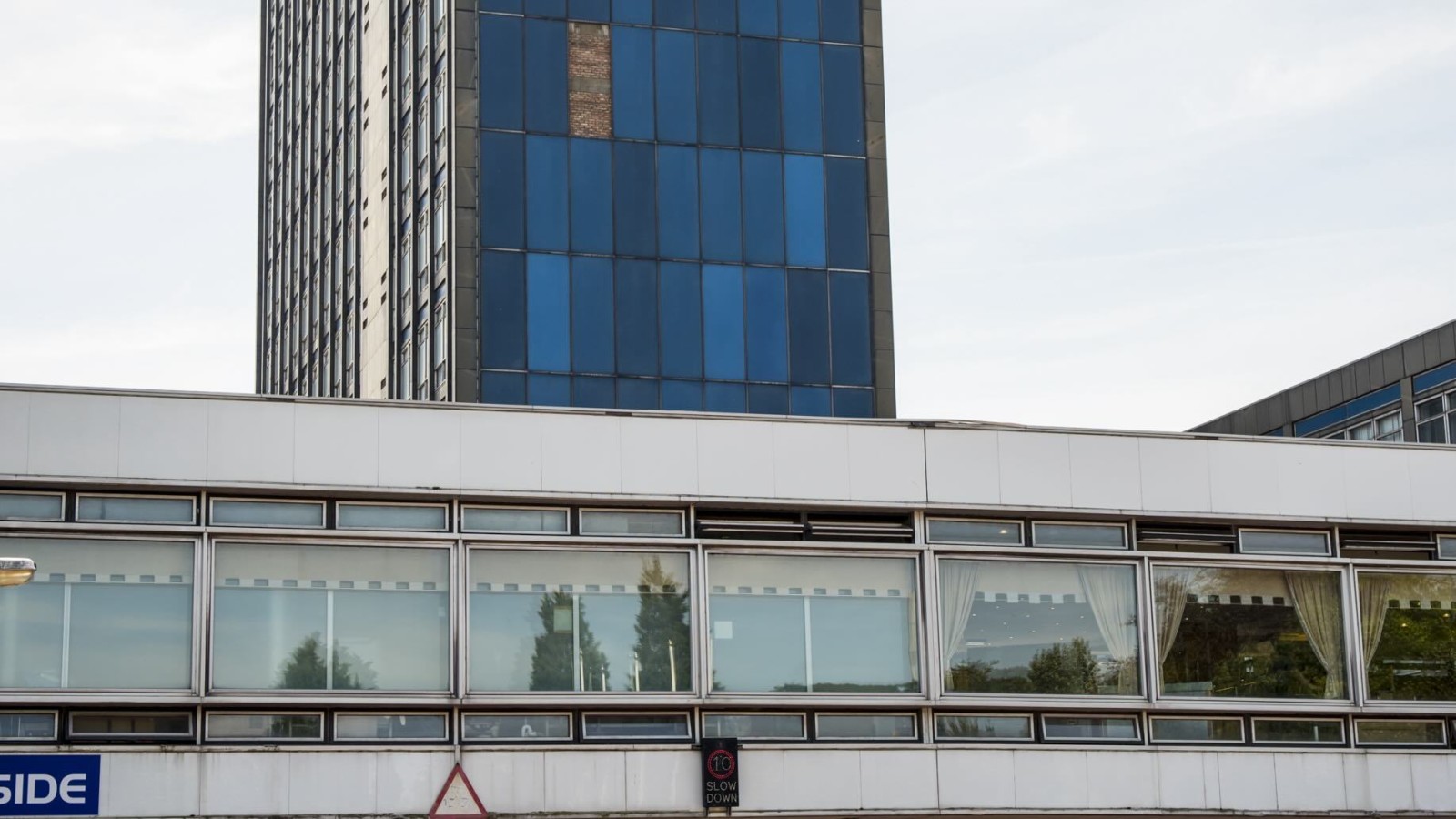 A large tower building stands in daylight against a blue/grey sky. The building has one whole side of blue windows. In front of the tower is a covered walkway over a car park. This is the former Pilkington's Glass Headquarters at Alexandra Park.