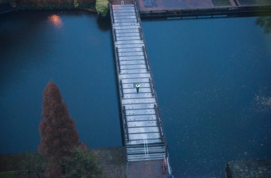 An aerial view of a person in a high visibility jacket walking across a bridge in evening light. The bridge crosses a body of dark, navy water, reflecting the evening sky.