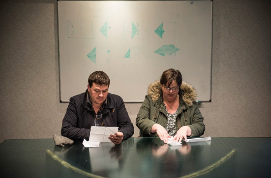 Two people sit at a table wearing coats. One of them reads from a piece of paper and the other is folding a piece of paper.