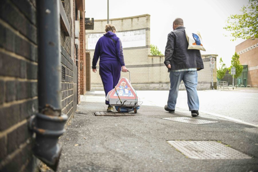 Two people walk, with their backs to us. They are walking on pavement next to a brick wall. One person, in a black leather jacket holds a canvas bag over their shoulder. The other in a purple tracksuit pulls a trolley which holds red triangular signs.