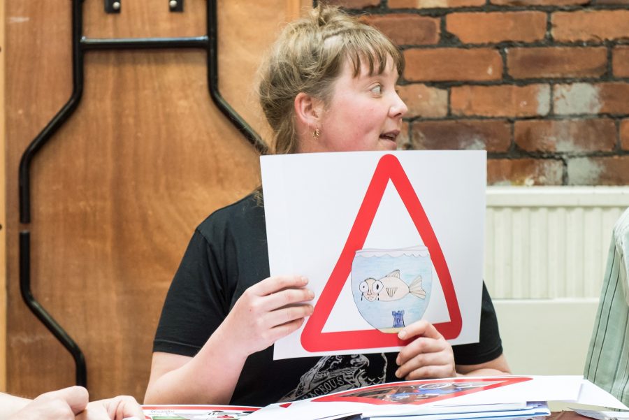 A person sits in a room with a brick wall and folded trestle behind them. They hold up a large piece of paper with a red triangular sign on it. On the table in front of them are many pieces of paper with drawing and more signs.