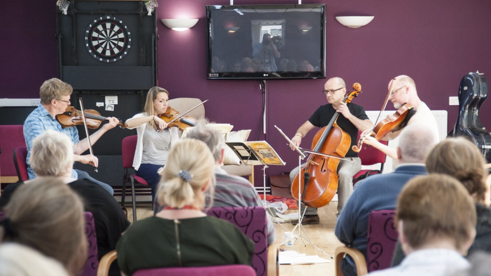 In a large room with purple walls, a dartboard and a TV on the wall sit 4 people playing instruments. Two playing violin and two playing cello. In front of them sit a number of people watching the musicians, with their backs to the camera.