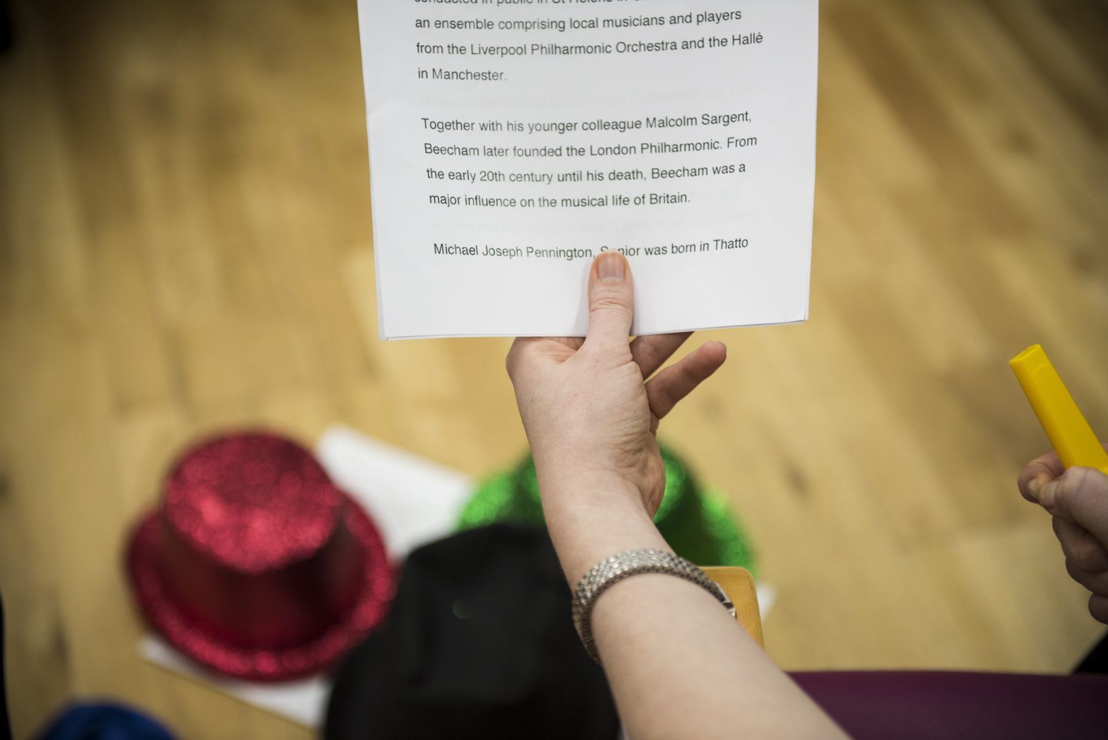 A hand holds a piece of paper with black text on, in the background a red and a green glittery hat sit on the floor.