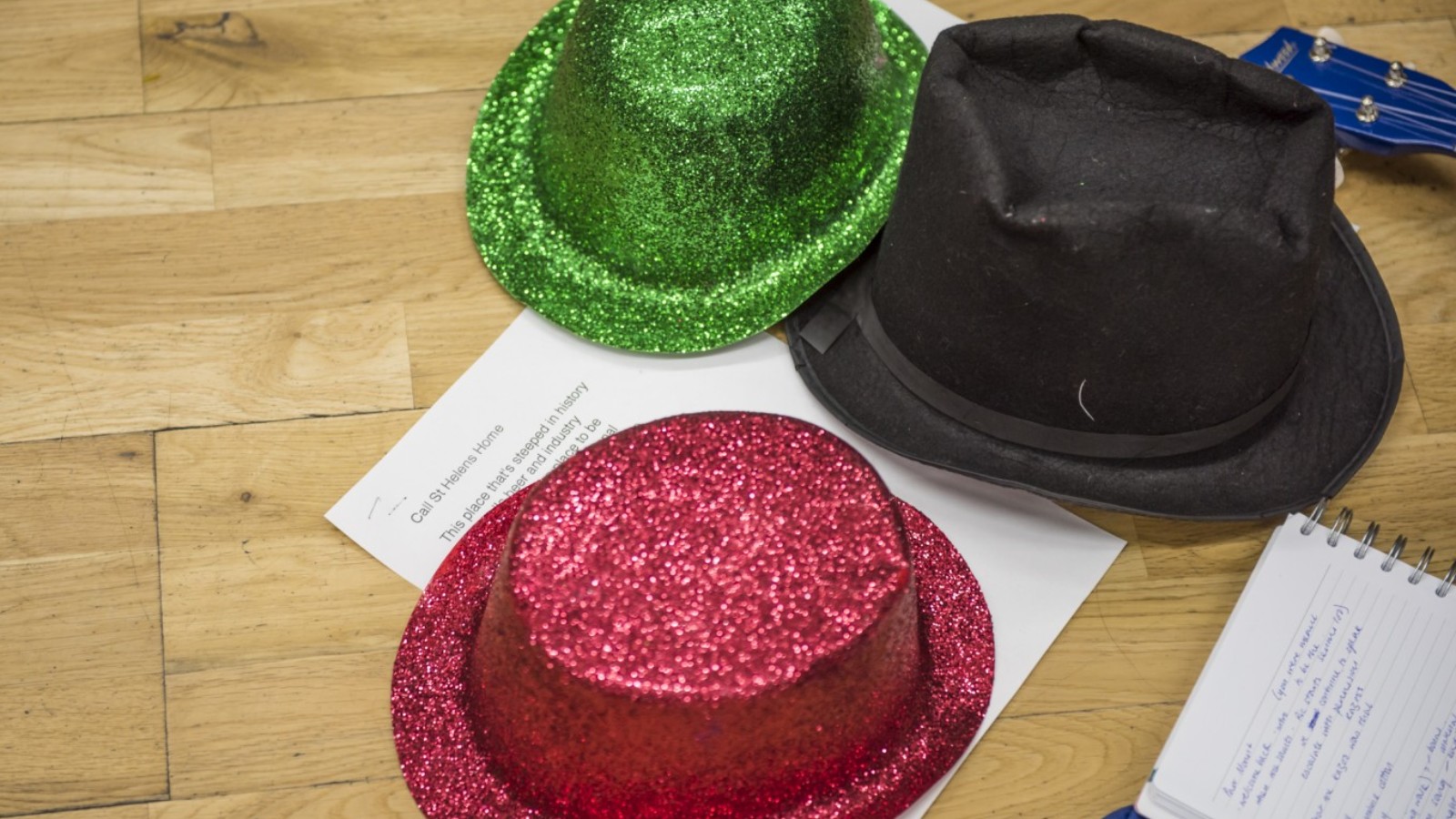 A red and a green glittery hat and a black top hate sit on a wooden floor with notepads and papers.