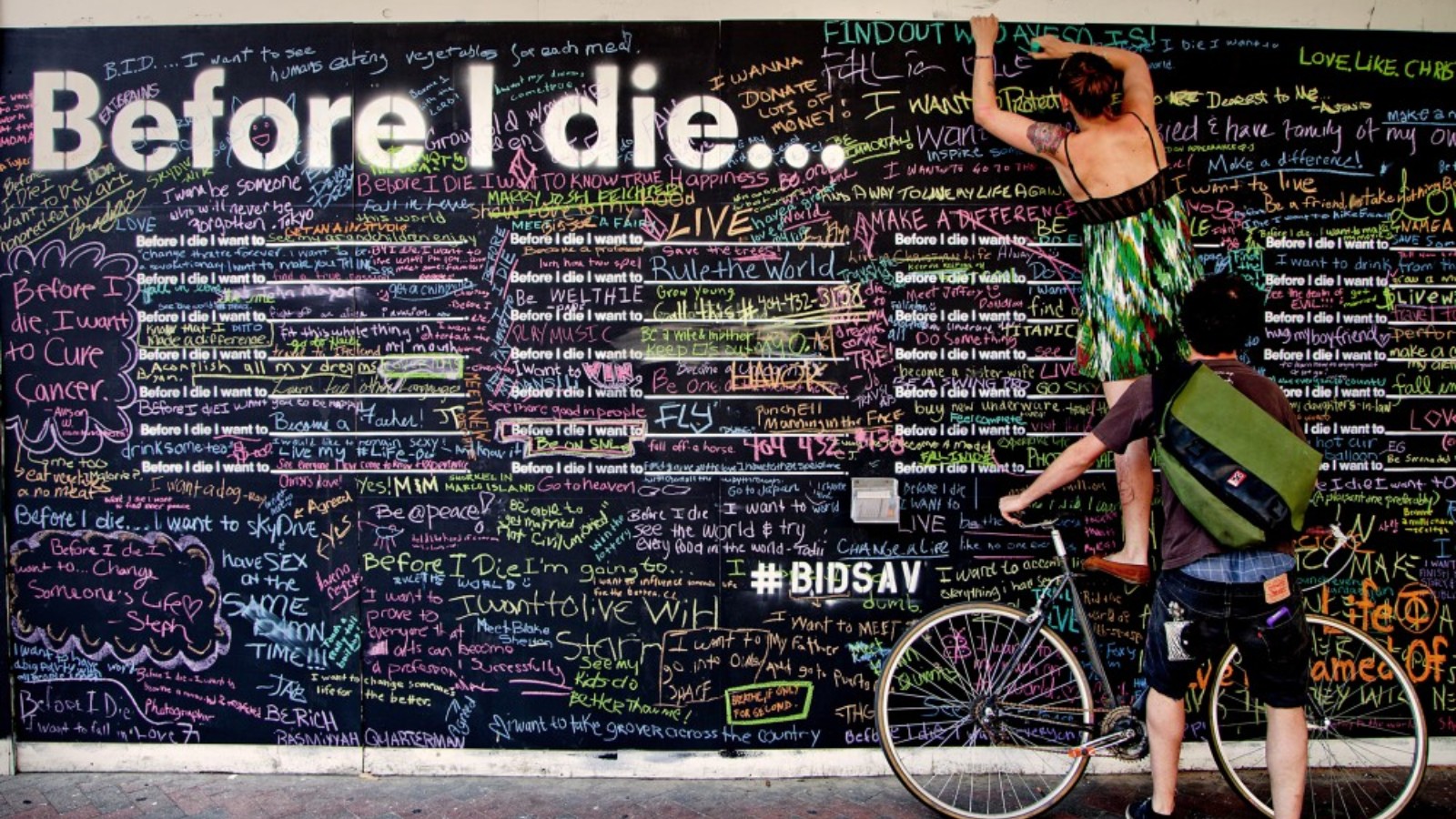 A blackboard reads 'Before I die...' in large white letters on the top left hand corner. The blackboard is covered in writing in many different colours of chalk. On the right there is a person wearing shorts and a t-shirt holding a bike against the blackboard while another person wears a green dress stands on the bike reaching to the top of the blackboard to write something.