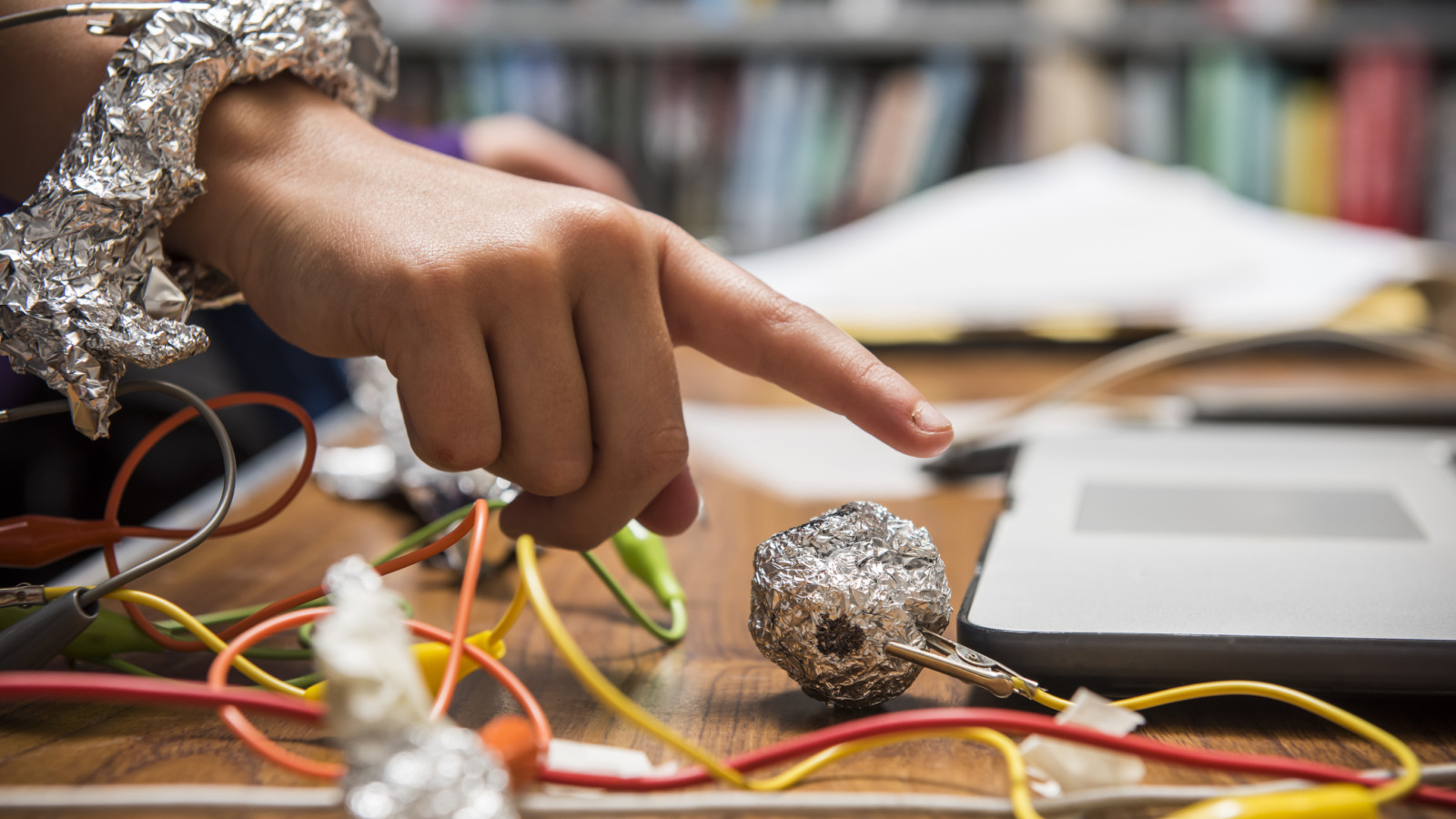 A young persons has foil wrapped around their wrist and are hovering their finger over a ball of foil. The pieces of foil are connected with yellow and red wires. There is a laptop computer in front of them.