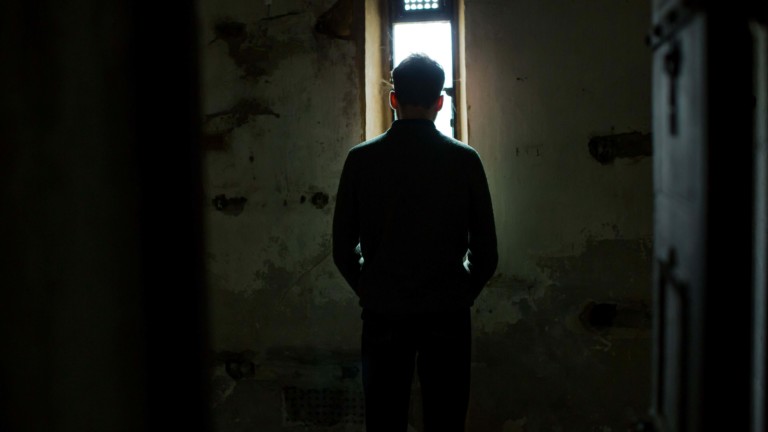 A person stands in front of a tall, thin window with their back to us. The light shines in through the window and the person is a silhouette, surrounded by bright light. We are looking at them though a doorway. The walls around the window are cracked and damaged.